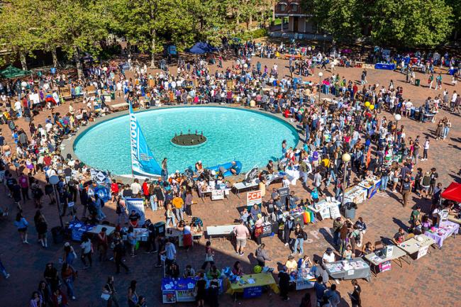 A large student event is set up outside on campus with two concentric circles of tables surrounding a large fountain. 